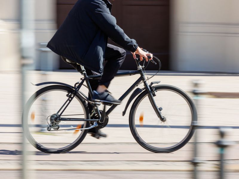 Person wearing dark clothes rides a bicycle quickly down a city street; background is blurred.