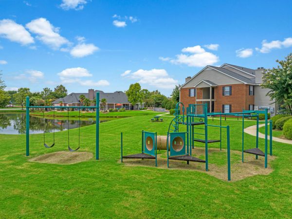 Playground with swings and climbing equipment on green lawn near a pond and apartment buildings under a blue sky.