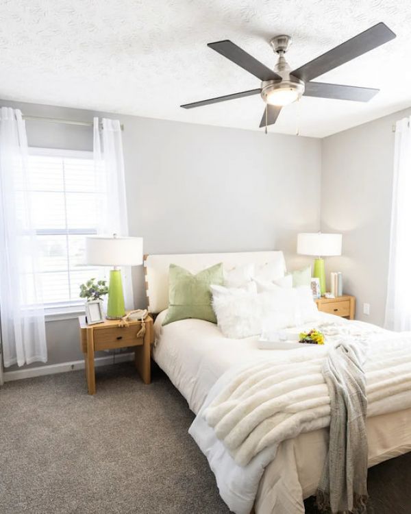 Bright bedroom with a neatly made bed, green pillows, bedside tables, and large windows with sheer curtains.