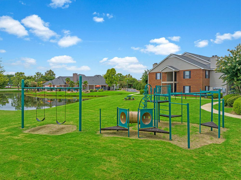 Playground with swings and climbing equipment on green lawn near a pond and apartment buildings under a blue sky.