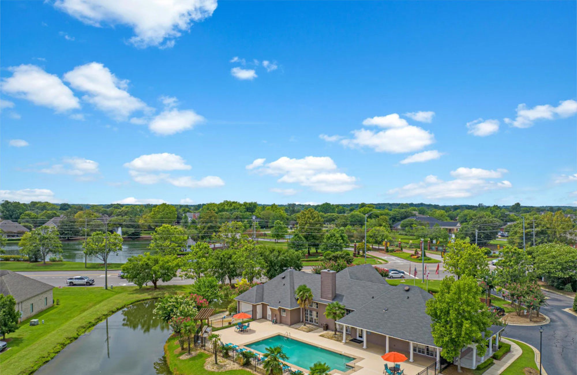 Aerial view of a clubhouse with a pool, palm trees, and a pond in a suburban neighborhood on a sunny day.