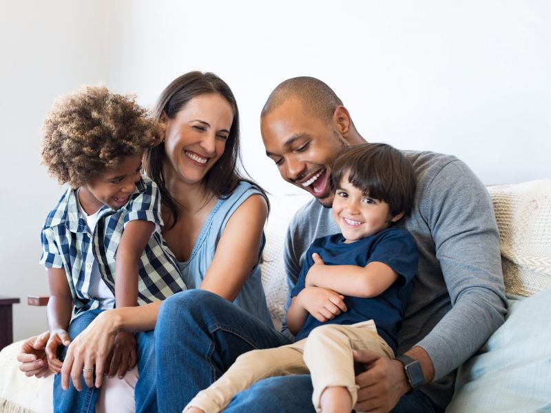 A happy family of four sits on a couch, smiling and laughing together.