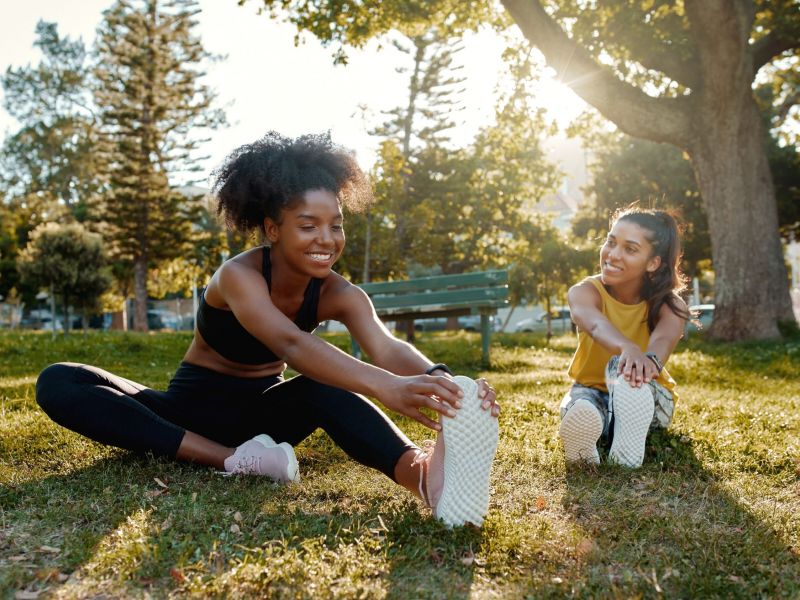 Two women sitting on grass, stretching their legs, and smiling in a sunny park with trees in the background.