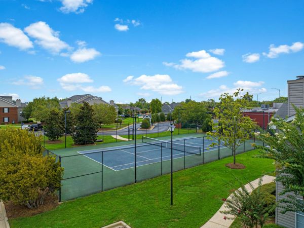 A fenced outdoor tennis court with trees and residential buildings in the background under a blue sky.