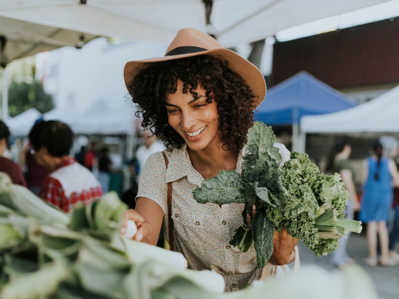 Smiling woman in a hat holding leafy greens at an outdoor market stall.