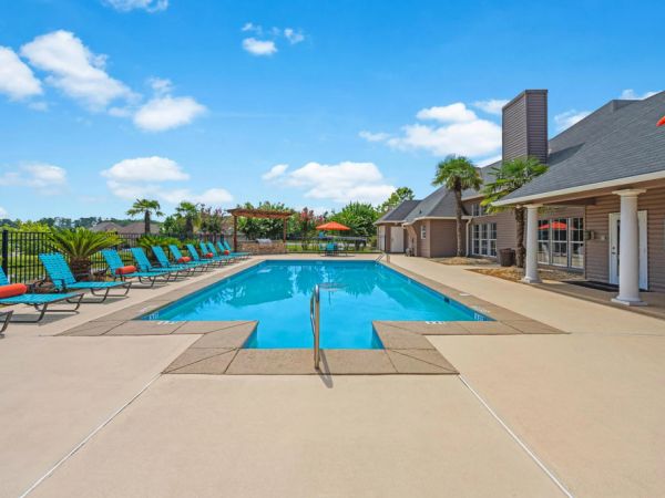 Outdoor swimming pool with lounge chairs, umbrellas, and a clubhouse under a bright blue sky.