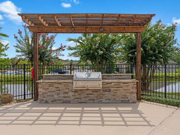Outdoor grill with a stone base and wooden pergola, set on a patio near a black metal fence and pond.